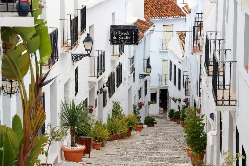 A narrow, cobblestone street in Frigiliana, Andalusia, Spain, lined with whitewashed buildings, black wrought-iron balconies, potted plants, and a sign for "La Parada del Zacatín."